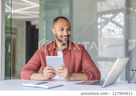 A smiling businessman sits at his desk, holding a tablet while working with a laptop in a modern office environment. Focus is on his expression. A smiling businessman sits at his desk, holding a tablet while working with a laptop in a modern office environment. Focus is on his expression. 127904811