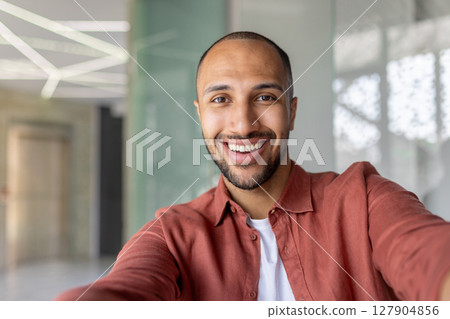 A close-up shot captures a smiling man in a casual orange shirt, taking a selfie indoors with a bright, welcoming expression. A close-up shot captures a smiling man in a casual orange shirt, taking a selfie indoors with a bright, welcoming expression. 127904856