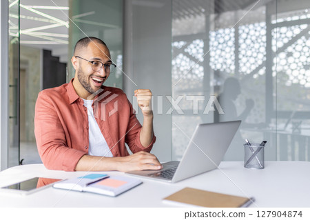 A joyful man celebrates success while working on a laptop. His elation is evident with fist raised, showcasing his excitement. 127904874