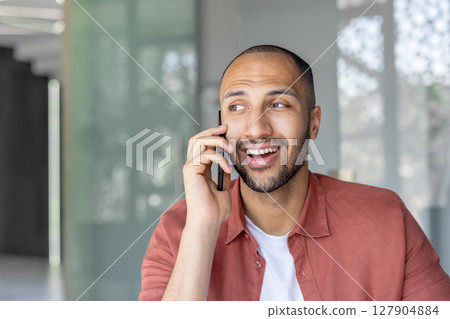A smiling man in a red shirt is on a call, appearing engaged and looking away with a happy expression. 127904884