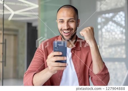 Excited man looking at his phone screen with a raised fist, celebrating success with a smile. Excited man looking at his phone screen with a raised fist, celebrating success with a smile. 127904888