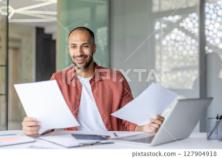 Successful businessman doing paperwork, man in casual clothes checking documents and bills, satisfied with results of activity smiling, working inside office with laptop. Successful businessman doing paperwork, man in casual clothes checking documents and bills, satisfied with results of activity smiling, working inside office with laptop. 127904918