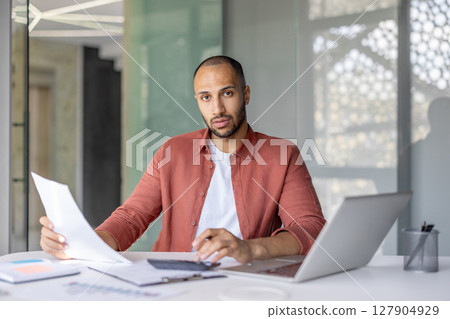 A focused businessman reviews paperwork at his desk, a laptop and calculator beside him, inside a bright office space. A focused businessman reviews paperwork at his desk, a laptop and calculator beside him, inside a bright office space. 127904929