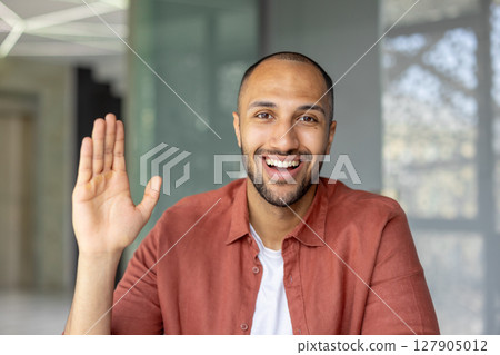 A happy man waves at the camera, smiling broadly and showing off his open palm. He's wearing a red-orange shirt. A happy man waves at the camera, smiling broadly and showing off his open palm. He's wearing a red-orange shirt. 127905012
