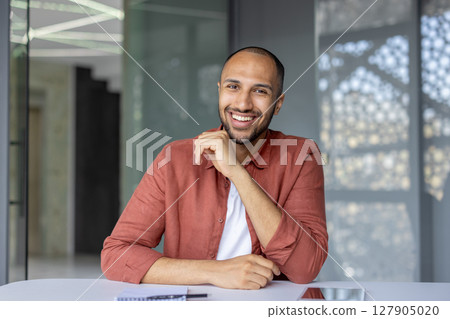 A handsome man is sitting at a desk in front of a window smiling. He is wearing a coral shirt over a white t-shirt and has a beard A handsome man is sitting at a desk in front of a window smiling. He is wearing a coral shirt over a white t-shirt and has a beard 127905020