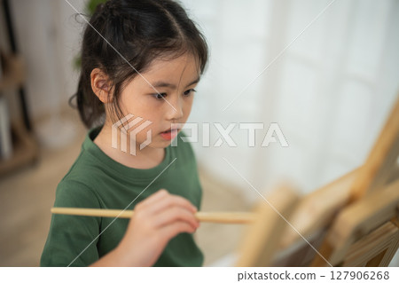 Young girl focused on painting at art easel with paintbrush in hand, showcasing creativity and imagination in a bright and inspiring environment 127906268