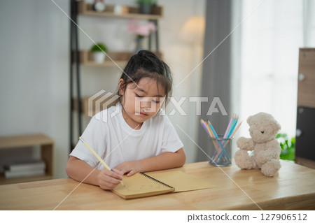 Young girl engaged in creative writing or drawing in a cozy indoor setting with soft light, surrounded by a teddy bear and colorful art supplies on a wooden table 127906512