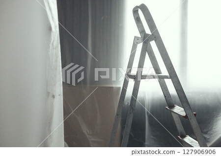 Sunlight filters through sheer curtains, casting a soft glow on a metal ladder in a minimalist room. The scene evokes themes of renovation and simplicity in a serene space. 127906906