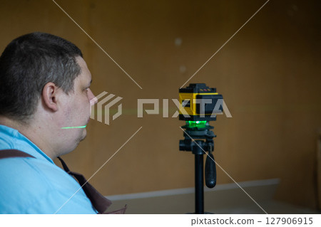 A man takes measurements and aligns using a laser level on a tripod. The green beam of the laser level provides precise horizontal alignment for interior construction and renovation work. A man takes measurements and aligns using a laser level on a tripod. The green beam of the laser level provides precise horizontal alignment for interior construction and renovation work. 127906915