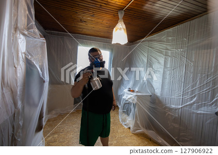 A male painter wearing a protective mask and using a paint sprayer repairs a room covered with plastic sheeting for protection during home renovation. A man is doing renovation work in his home. 127906922
