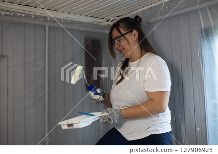 Woman in a white shirt and safety goggles painting a room with a roller. She holds a paint tray and wears gloves, surrounded by plastic sheeting, creating a diy home improvement scene. 127906923