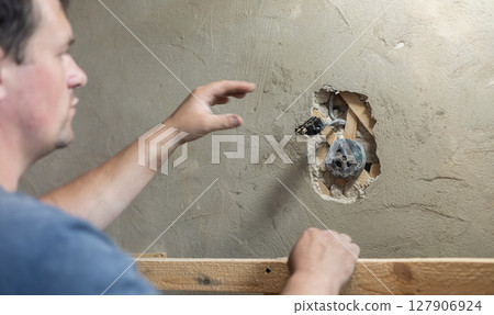 Electrician carefully positioning a new electrical socket into a damaged wall section, demonstrating the process of electrical installation during a home renovation project. Electrician carefully positioning a new electrical socket into a damaged wall section, demonstrating the process of electrical installation during a home renovation project. 127906924