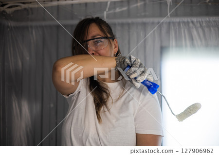 A female painter tiredly wipes her face while painting a ceiling. The female painter, wearing work gloves and safety glasses, holds a paint roller, indicating that she is painting. A female painter tiredly wipes her face while painting a ceiling. The female painter, wearing work gloves and safety glasses, holds a paint roller, indicating that she is painting. 127906962
