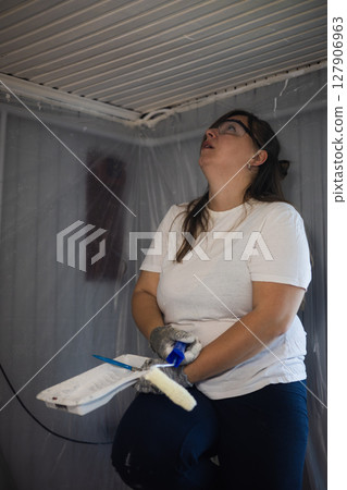 A woman in protective gear paints a ceiling with a roller, surrounded by plastic sheeting. The female painter is working on a home renovation project, focusing on improving the interior space. A woman in protective gear paints a ceiling with a roller, surrounded by plastic sheeting. The female painter is working on a home renovation project, focusing on improving the interior space. 127906963