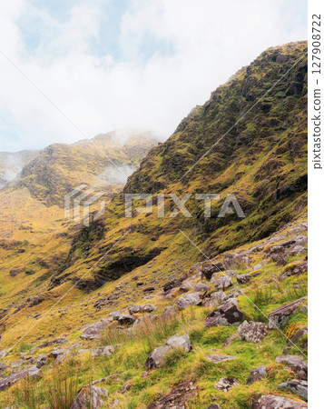 Rugged mountain terrain of Carrauntoohil in County Kerry, Ireland. Walkway trails and rocky landscape are covered with green and golden vegetation. Clouds are present in the sky. Rugged mountain terrain of Carrauntoohil in County Kerry, Ireland. Walkway trails and rocky landscape are covered with green and golden vegetation. Clouds are present in the sky. 127908722