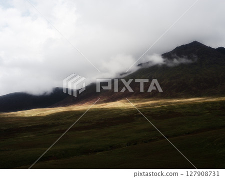 Beautiful view of Carrauntoohill mountain under a cloudy sky. Light shines across the green fields at the base of the mountain. Clouds cover the peak of the rugged, rocky terrain. 127908731