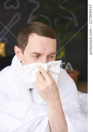 Caucasian young male sneezing into tissue under white blanket indoors 127909437