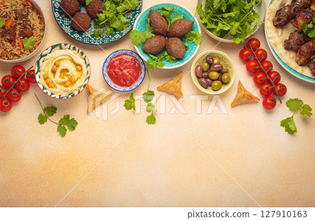Top view of a vibrant table filled with lebanese dishes and mezze, featuring meat kebabs, falafel, kibbeh, hummus, sambousek, rice, olives. Middle eastern or Arab dishes, space for text Top view of a vibrant table filled with lebanese dishes and mezze, featuring meat kebabs, falafel, kibbeh, hummus, sambousek, rice, olives. Middle eastern or Arab dishes, space for text 127910163