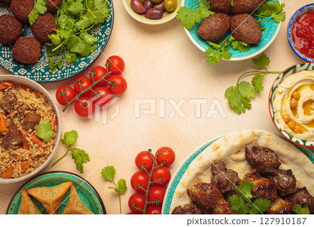 Top view of a vibrant table filled with lebanese dishes and mezze, featuring meat kebabs, falafel, kibbeh, hummus, sambousek, rice, olives. Middle eastern or Arab dishes, space for text Top view of a vibrant table filled with lebanese dishes and mezze, featuring meat kebabs, falafel, kibbeh, hummus, sambousek, rice, olives. Middle eastern or Arab dishes, space for text 127910187