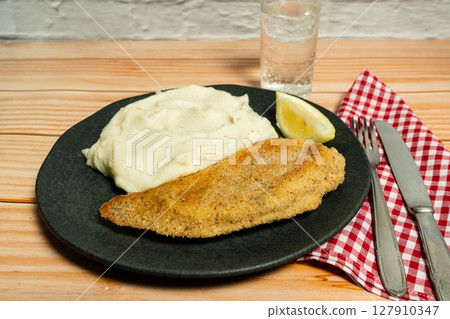 Breaded fish fillet with mashed potatoes and lemon slice on black plate Breaded fish fillet with mashed potatoes and lemon slice on black plate 127910347