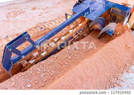 Heavy trencher equipment grinds through soil while preparing trench for underground utilities in an active construction zone Heavy trencher equipment grinds through soil while preparing trench for underground utilities in an active construction zone 127910492