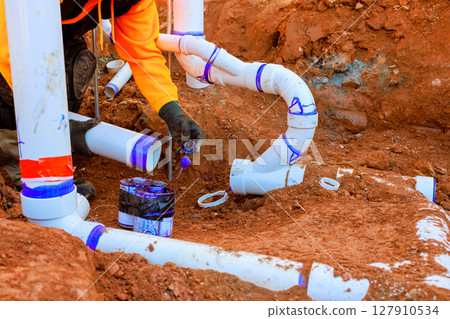 Construction worker in an orange jacket installs drainage sewage PVC pipes in trench, ensuring proper alignment. 127910534