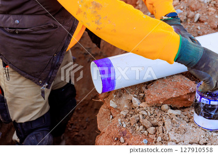 Worker is preparing section of PVC pipe with adhesive in trench for plumbing installation at construction site. 127910558
