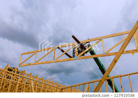 Construction crew operates machinery to raise wooden beams into place amidst overcast skies at building site. 127910586