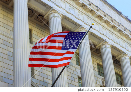 An American flag waves in front of grand government structure featuring tall columns stately architecture. An American flag waves in front of grand government structure featuring tall columns stately architecture. 127910590