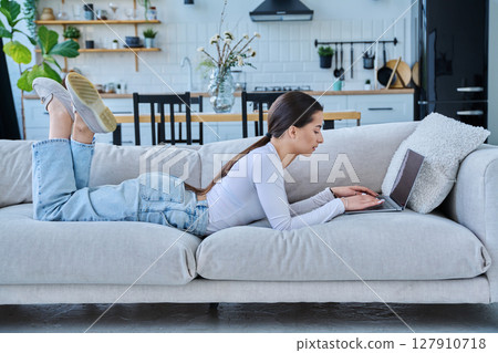 Portrait of young woman lying on sofa typing on laptop, in home interior Portrait of young woman lying on sofa typing on laptop, in home interior 127910718