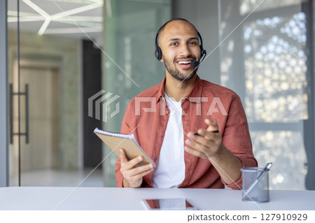 Webcam view, man with headset smiling and looking at camera holding notebook in hands. Company employee at online business conference meeting. 127910929