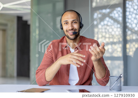 A smiling man wearing a headset gestures during a video conference, sitting at a desk indoors, communicating. 127910930