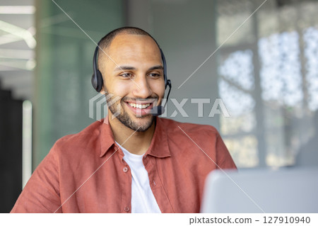 A smiling man wearing a headset appears to be in a customer service or call center environment, possibly assisting a client. A smiling man wearing a headset appears to be in a customer service or call center environment, possibly assisting a client. 127910940