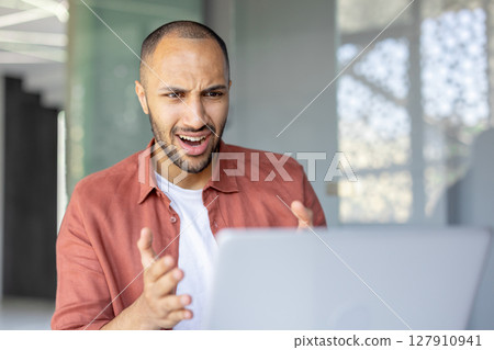 A young man looks surprised at his laptop during a video call in a modern, bright room. He is reacting to something on the screen A young man looks surprised at his laptop during a video call in a modern, bright room. He is reacting to something on the screen 127910941