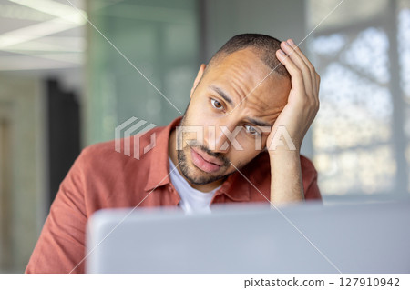 Sad anxious man working with laptop inside office. Close-up of office worker in depression holding hand behind head, looking at laptop screen suffering despair. Sad anxious man working with laptop inside office. Close-up of office worker in depression holding hand behind head, looking at laptop screen suffering despair. 127910942