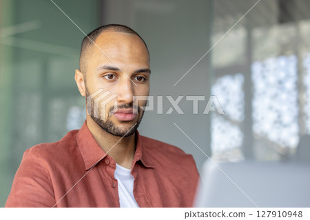 A focused, young man with a beard looks at a laptop screen, in a workspace with natural light filtering through the background. 127910948