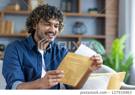 A cheerful man opening an envelope, appearing excited and happy. He's wearing a denim shirt in a comfortable setting. 127910963