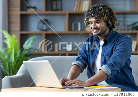 A smiling man with curly hair wearing a headset works on a laptop while sitting on a sofa at home. 127911005