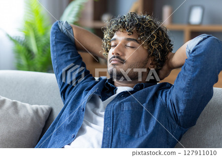 A relaxed man with curly hair sits on a couch indoors with his eyes closed and hands behind his head, enjoying a moment of tranquility. 127911010