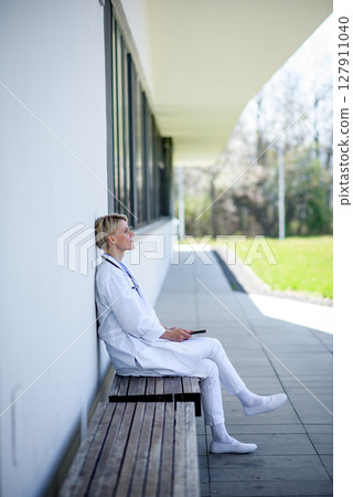 Young female doctor taking a break, sitting on bench in front of hospital. Young female doctor taking a break, sitting on bench in front of hospital. 127911040