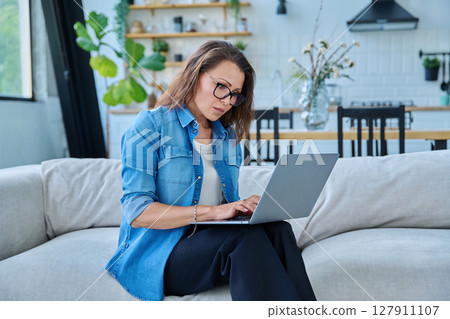 Portrait of middle aged woman using laptop sitting on sofa at home 127911107