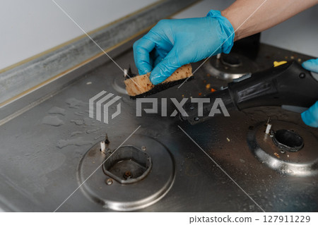 Homemaker male wearing blue glove cleaning dirty gas stove, removing grease and food remnants with sponge and detergent, close-up. Concept of household chores and kitchen hygiene. Homemaker male wearing blue glove cleaning dirty gas stove, removing grease and food remnants with sponge and detergent, close-up. Concept of household chores and kitchen hygiene. 127911229