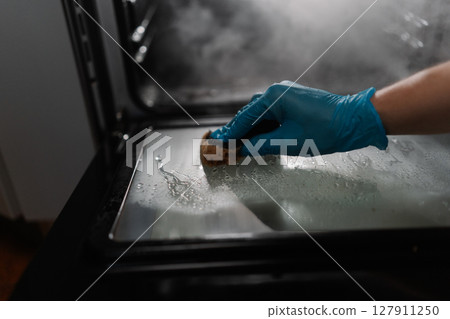 Close-up view of cleaning company worker wearing blue glove using sponge for cleaning glass door of gas oven in dark kitchen room. Concept of hygiene and cleanliness in domestic environment. 127911250