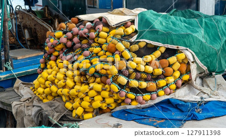 Weathered yellow and brown fishing floats and tangled nets piled on a dockside boat, with parts of the vessel and harbor scene in the background. 127911398