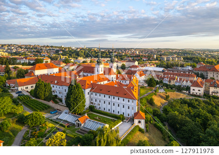 Strahov Monastery reveals its stunning architecture illuminated by the soft morning light. An aerial view showcases the vibrant red roofs and lush surroundings at dawn in Prague. 127911978