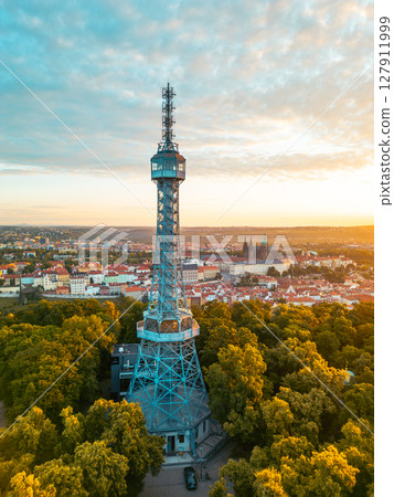 Petrin Lookout Tower rises above lush greenery as dawn breaks over Prague. The warm hues of sunrise illuminate the cityscape, showcasing Prague Castle in the distance. 127911999