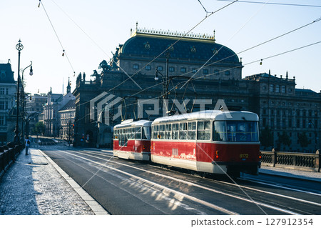 Two red trams travel across a bridge near the National Theatre in Prague on a sunny morning, showcasing the vibrant public transport system in this historic city. 127912354