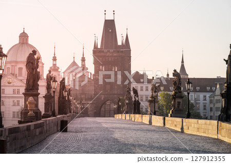 Early morning light bathes Charles Bridge in a golden hue, illuminating its historic statues and towers. The quiet Vltava river reflects the serene beauty of the scene. 127912355