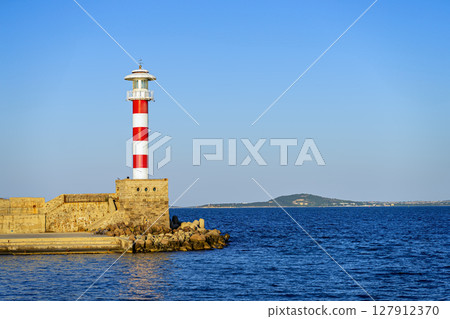 Tall lighthouse rises from stone pier with distant island and calm sea under clear sky. Concept of lighthouse, maritime stability, tranquil coastal Tall lighthouse rises from stone pier with distant island and calm sea under clear sky. Concept of lighthouse, maritime stability, tranquil coastal 127912370