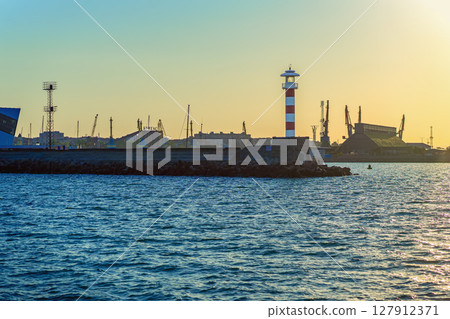 Industrial port with lighthouse at sunset. Lighthouse stands at edge of industrial port with cranes, golden sunset sky. Concept of lighthouse, urban harbor, contrast between nature and infrastructure Industrial port with lighthouse at sunset. Lighthouse stands at edge of industrial port with cranes, golden sunset sky. Concept of lighthouse, urban harbor, contrast between nature and infrastructure 127912371